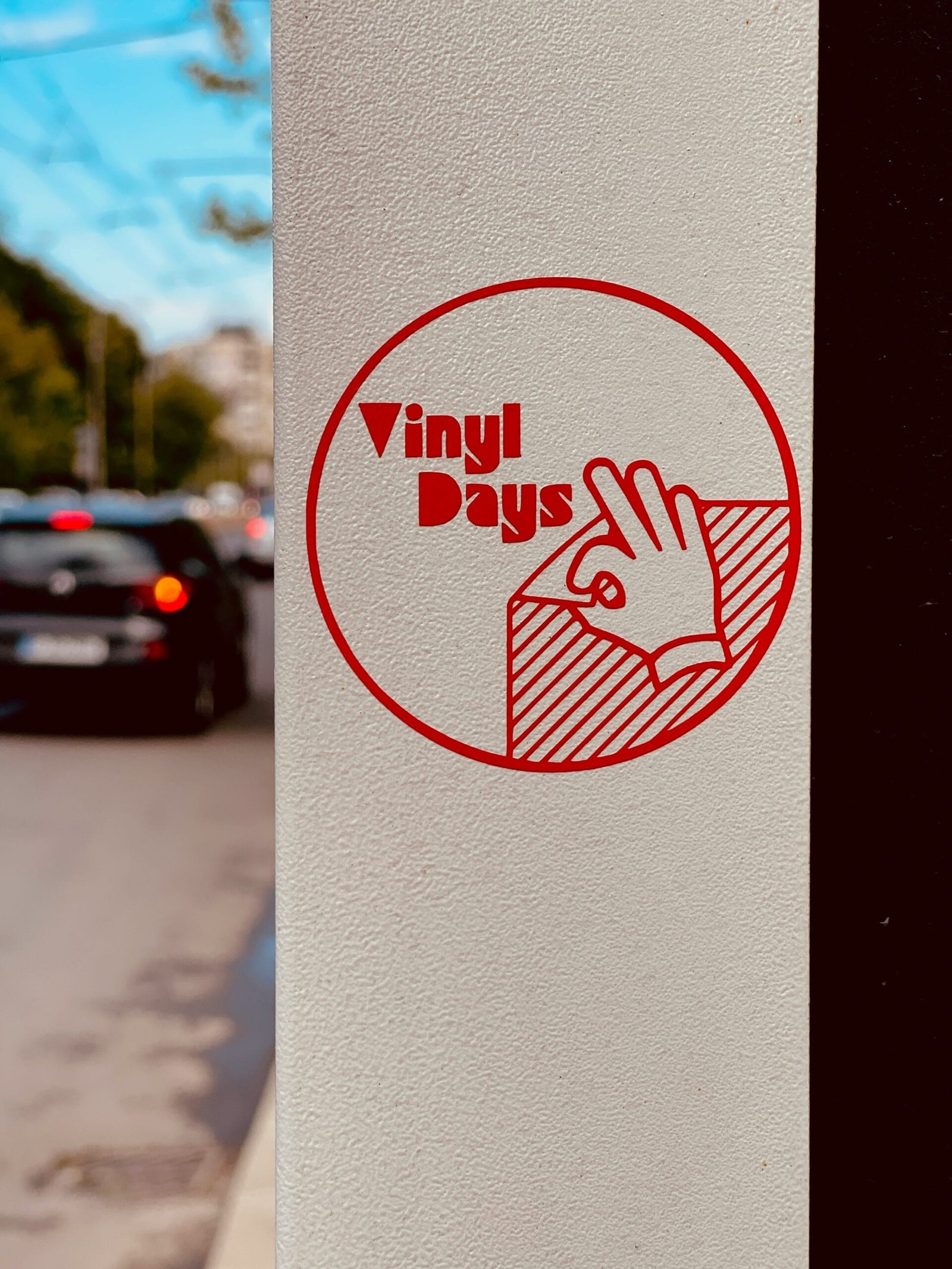 Vinyl Days Window Decal A red vinyl decal reading “Vinyl Days” with a hand gesture icon is applied to a light-colored window or wall, with a blurred street scene in the background.