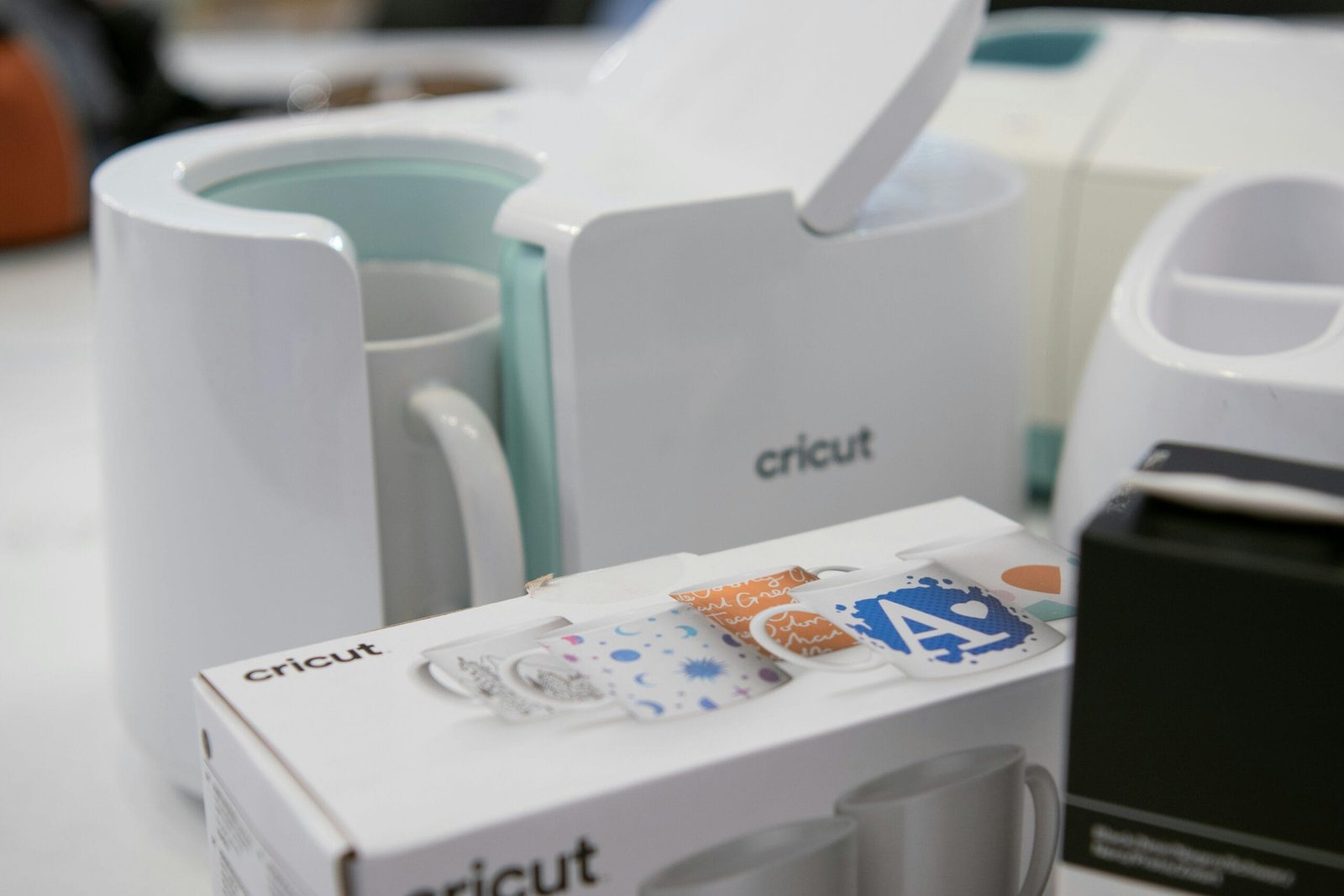 A Cricut mug press and accessories on a worktable, with a mug inside the press and a Cricut box in the foreground.